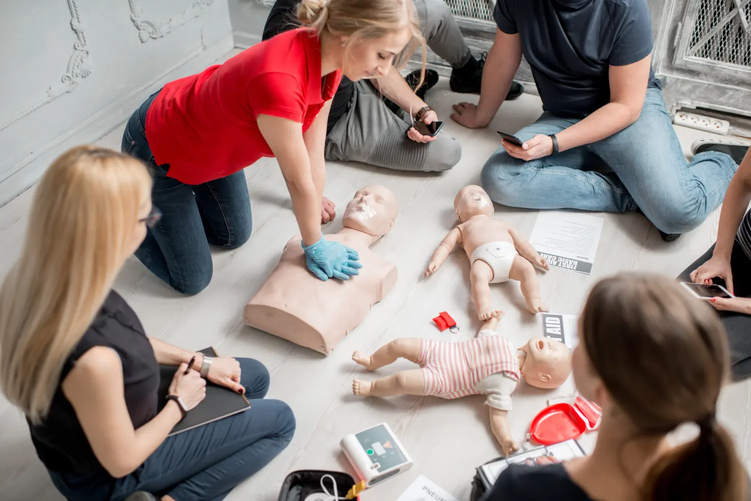 Eine junge Kursleiterin zeigt im Rahmen einer Erste-Hilfe-Gruppenschulung in Innenräumen, wie man mit einer Puppe Herzdruckmassagen durchführt.